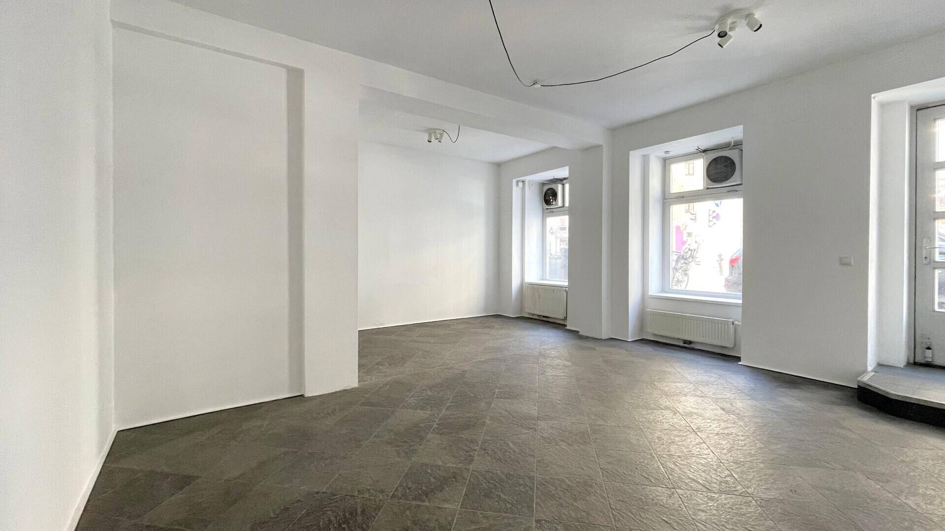 Heller, leerer Raum mit großen Fenstern zur Straße und grauen Fliesen, ideal für ein Geschäft oder Büro.
