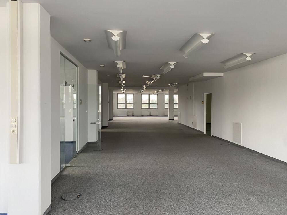 Geräumiger, leerer Büroraum mit grauem Teppichboden und vielen Fenstern, bereit zur Gestaltung.