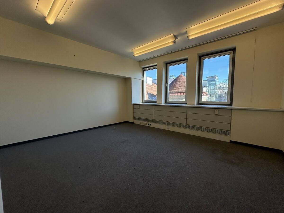 Großes, leeres Büro mit Teppichboden, hellgelben Wänden und drei Fenstern, die viel Tageslicht hereinlassen.
