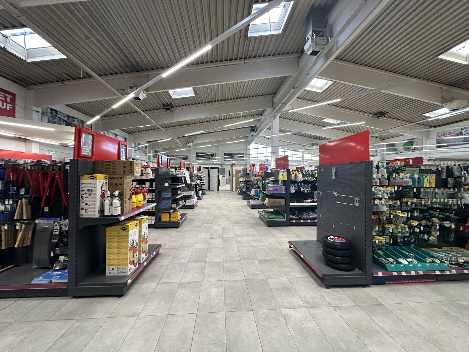 Langer Gang in einem Baumarkt mit zahlreichen Produkten in den Regalen und Tageslicht von oben.
