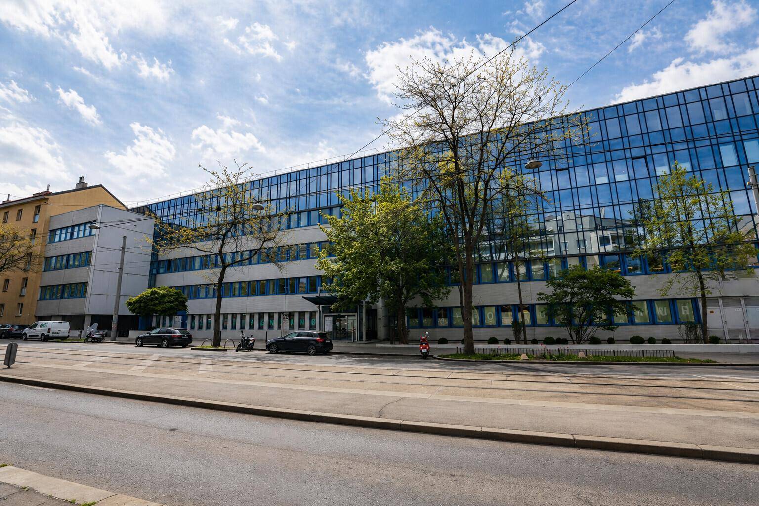 Lange Fassade eines modernen Bürogebäudes mit vielen Fenstern und Bäumen davor an einer Straße.