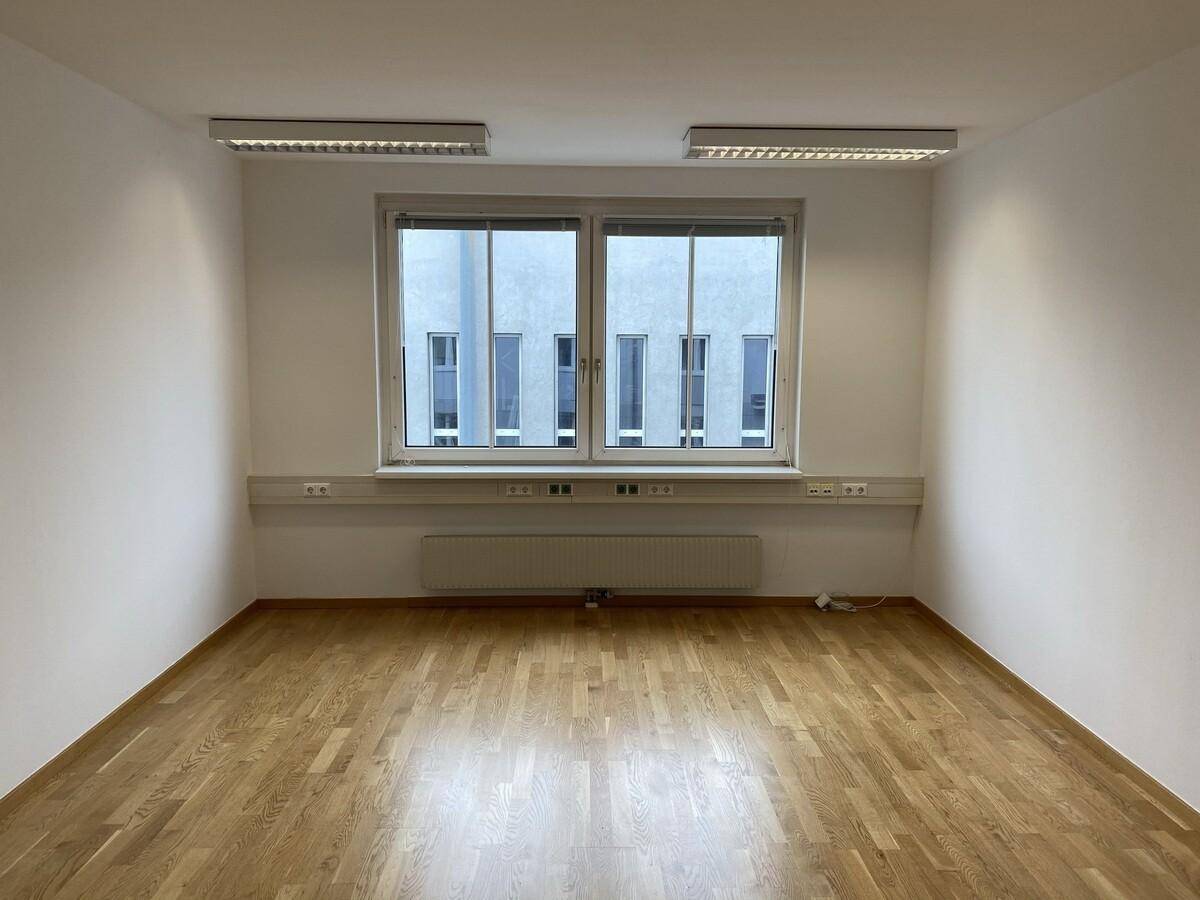 Leerer Büroraum mit Holzparkettboden, großen Fenstern und Blick auf ein gegenüberliegendes Gebäude.