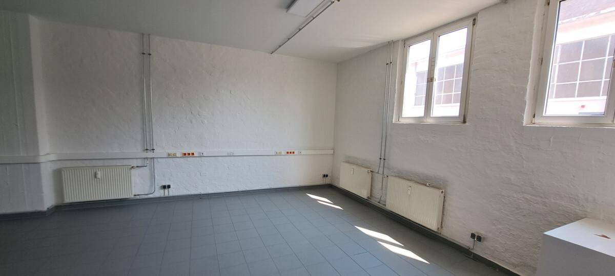 Heller, leerer Raum mit mehreren Fenstern, Heizkörpern und weißer Wandstruktur, ideal für Bürozwecke.