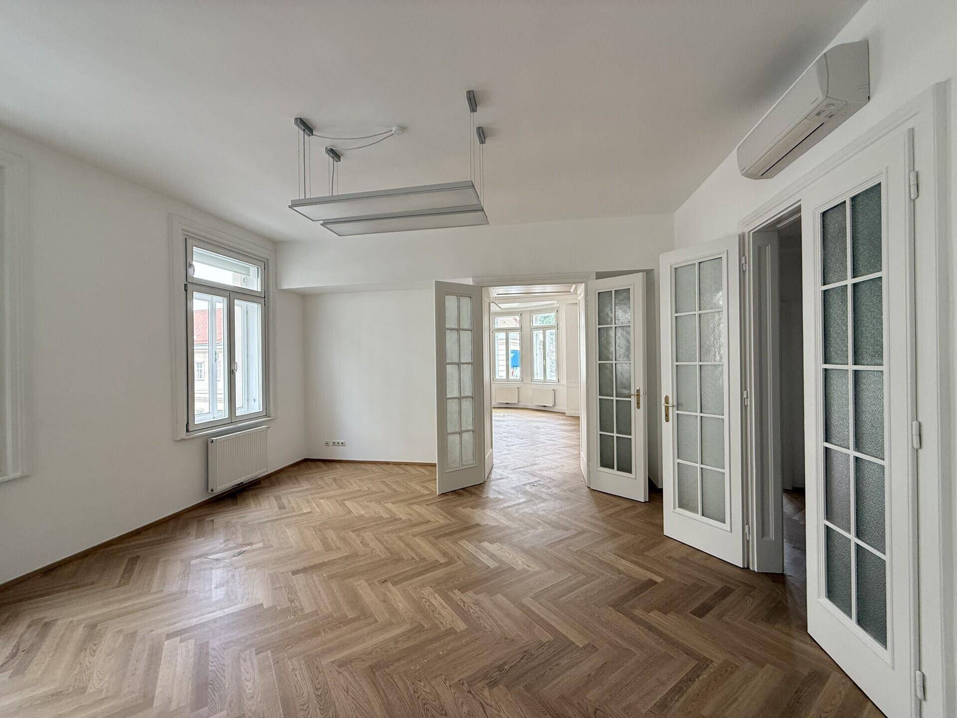 Helles Büro mit Fischgrätparkett, Flügeltüren und Klimaanlage an der Wand, bietet viel Tageslicht.
