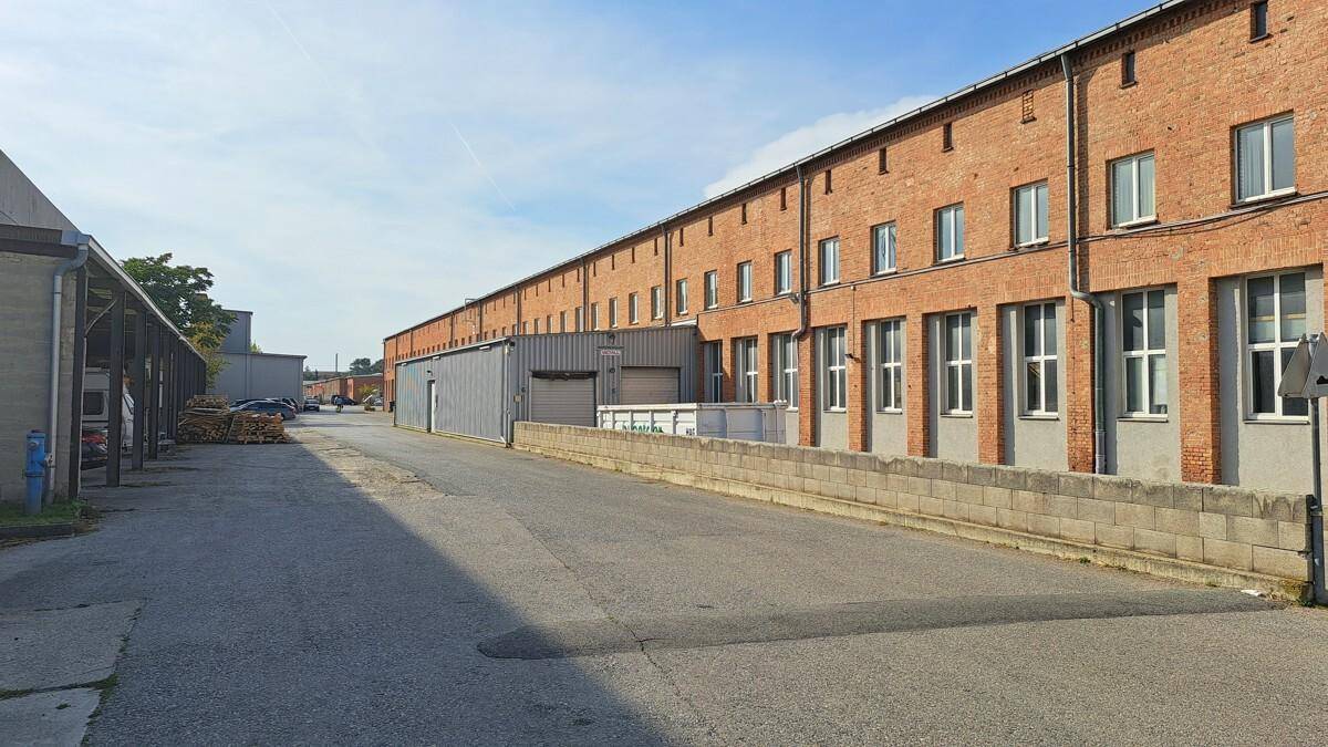 Lange Backsteinfassade eines Industriegebäudes mit vielen Fenstern und einem asphaltierten Zufahrtsweg.