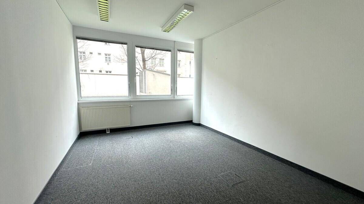 Heller, leerer Büroraum mit grauem Teppichboden, weißen Wänden und Heizkörpern unter den Fenstern.