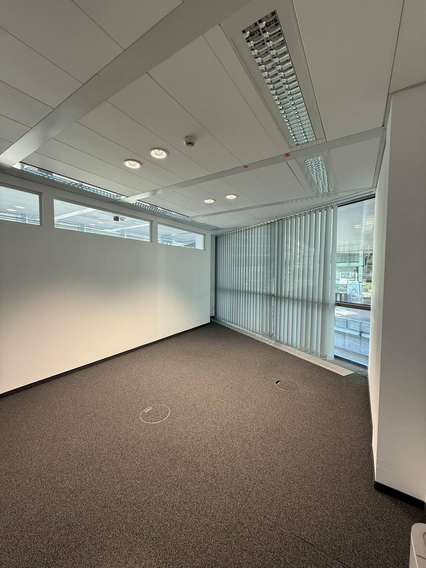 Unmöbliertes Büro mit Teppichboden, großen Fenstern und vertikalen Jalousien, die für Privatsphäre sorgen.