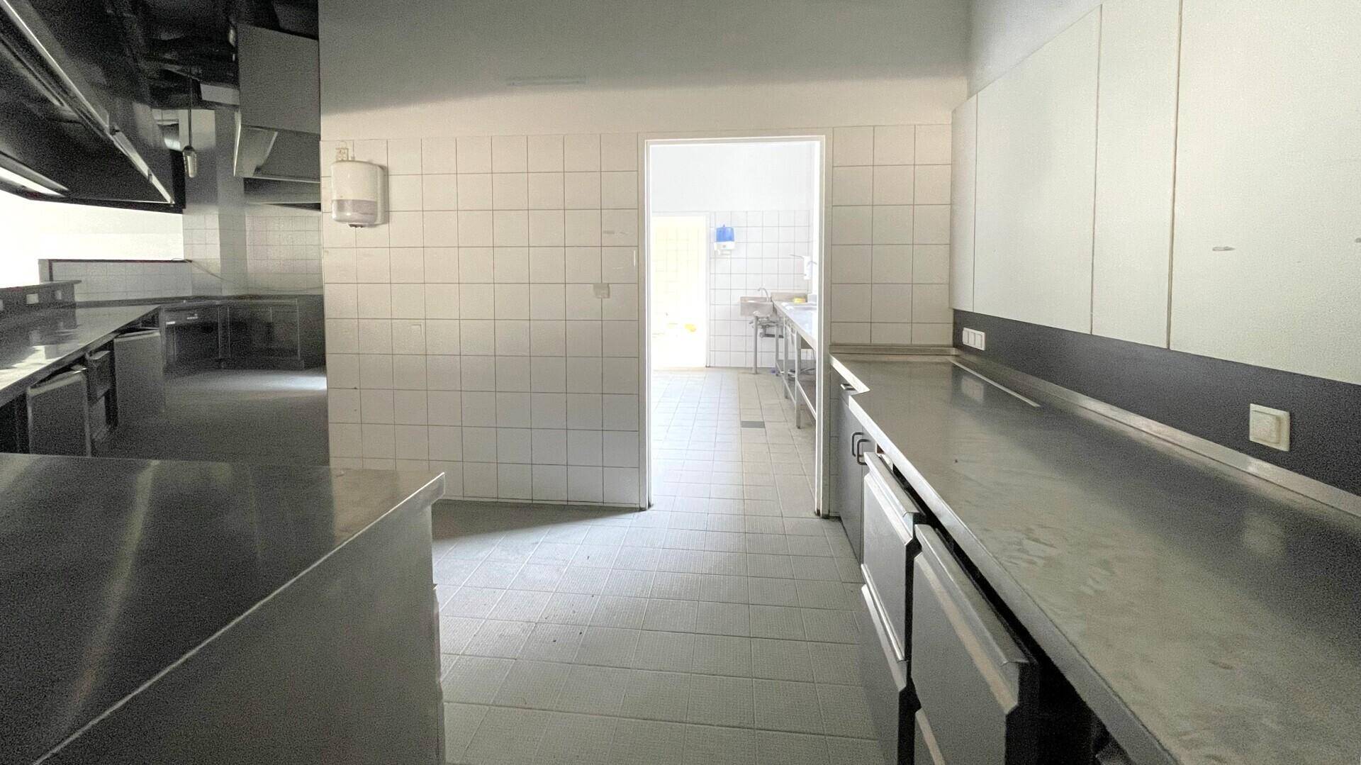 Gewerbeküche mit weißen Fliesenwänden, Edelstahlarbeitsflächen und Durchgang zu einem weiteren Raum.