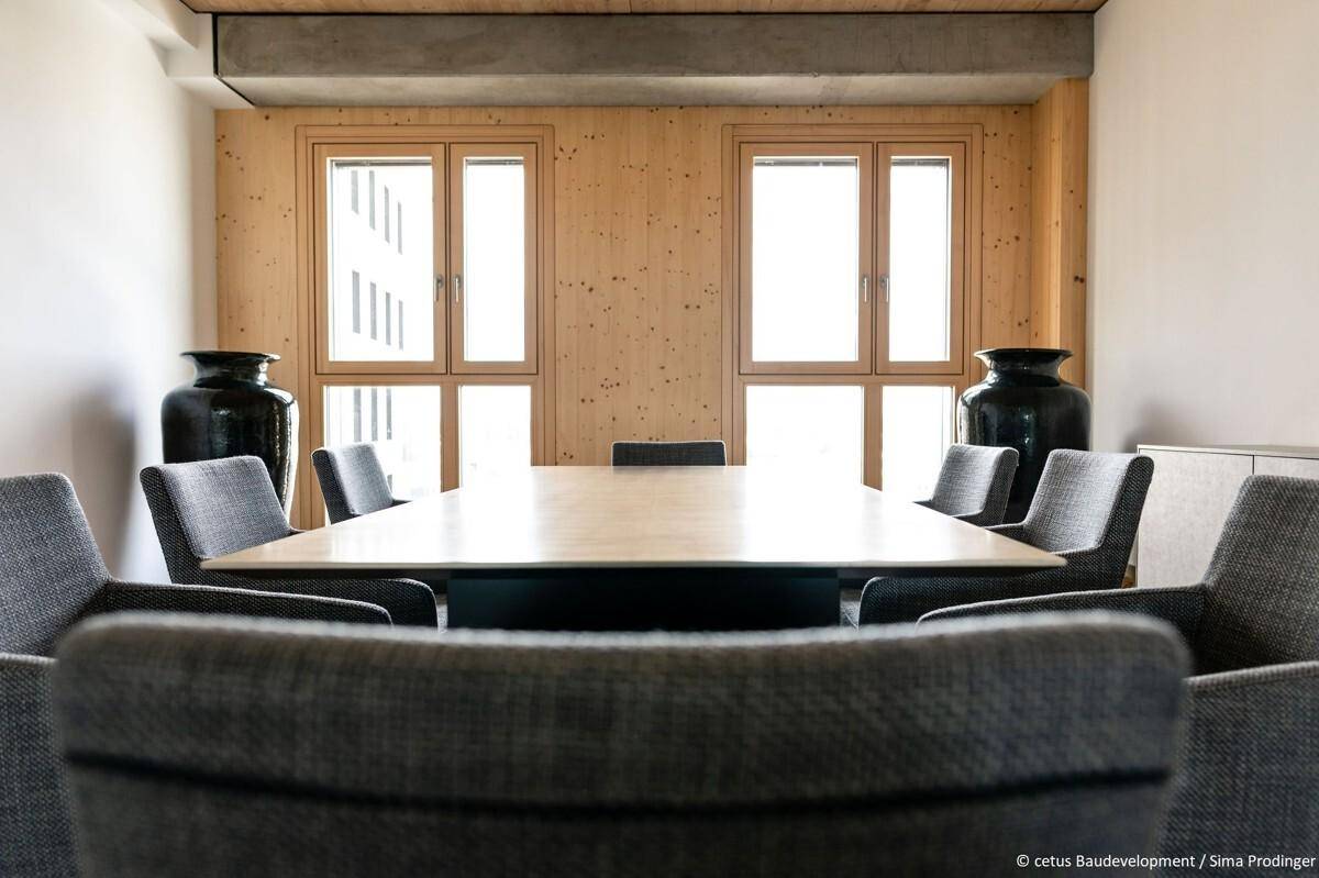 Moderner Konferenzraum mit großem Holztisch, grauen Stühlen und Holzwänden, hell beleuchtet.
