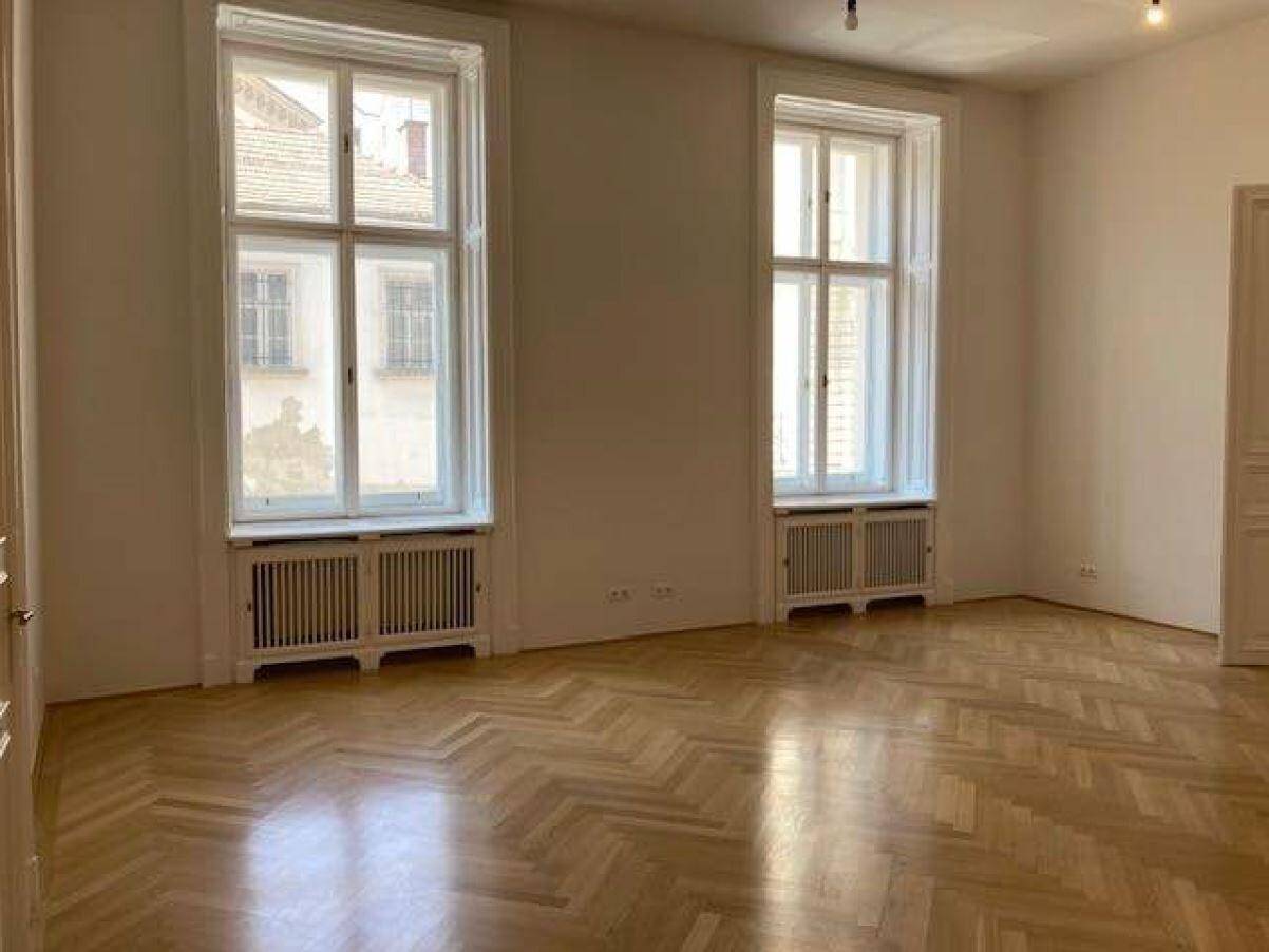 Geräumiges Zimmer mit hellem Fischgrätparkett, weißen Wänden und zwei hohen Fenstern.