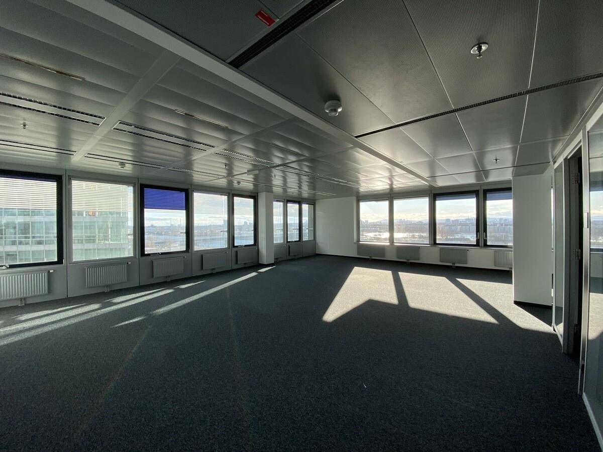 Großer, leerer Büroraum mit dunklem Teppichboden und vielen Fenstern, die einen weiten Stadtblick bieten.