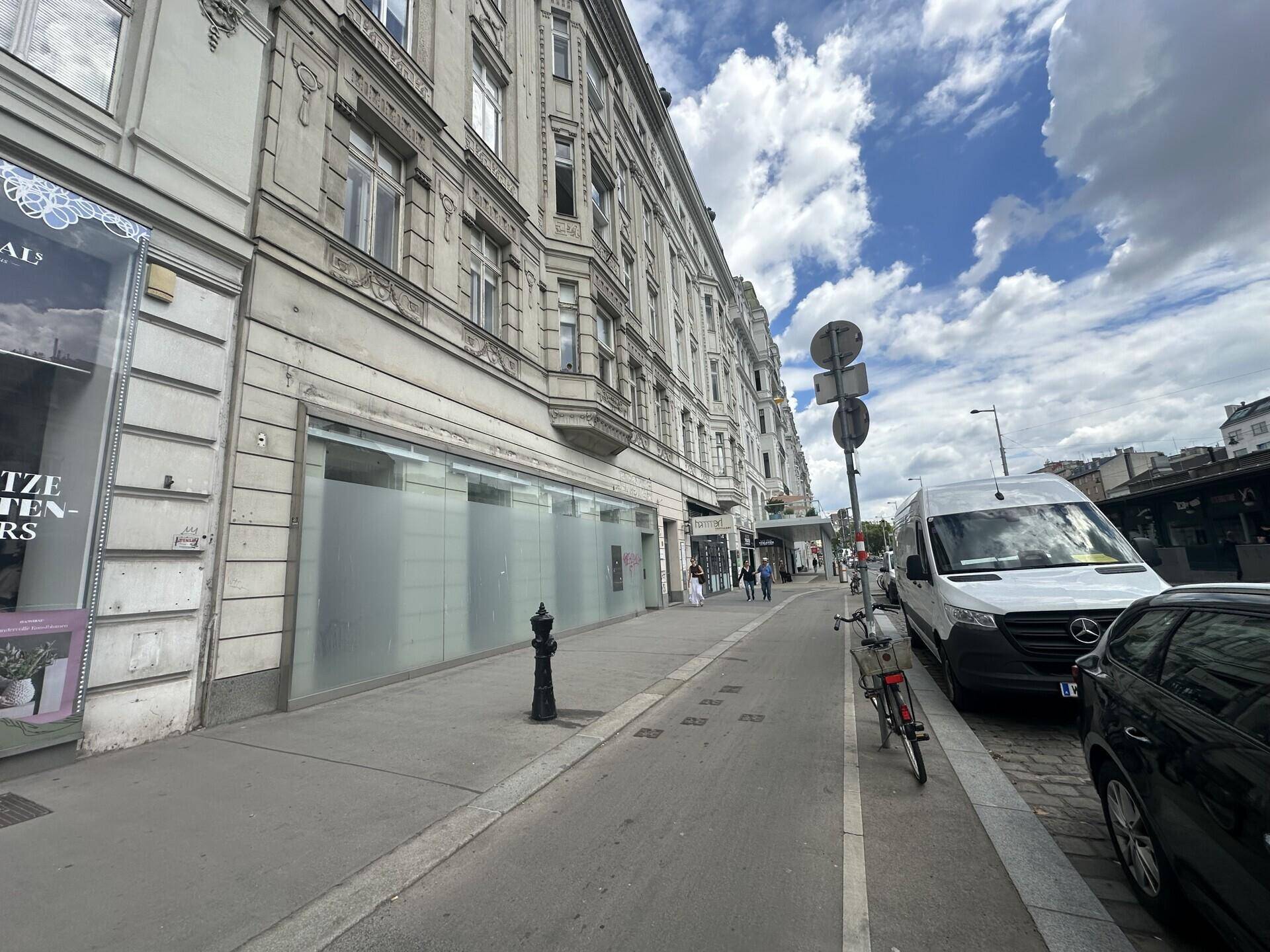 Seitenansicht des historischen Gebäudes mit Bürgersteig und parkenden Fahrzeugen unter bewölktem Himmel.