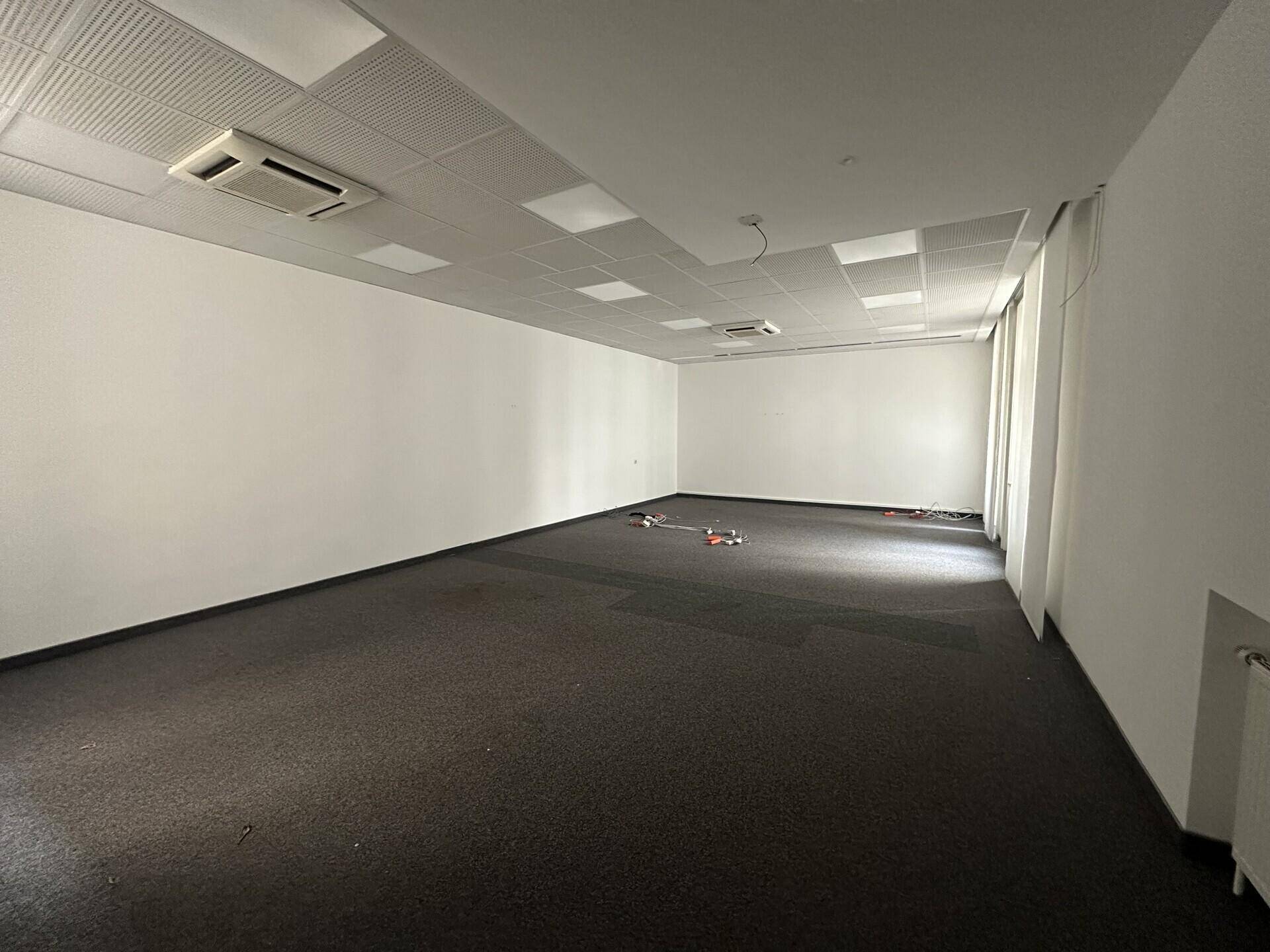 Leerer Büroraum mit Teppichboden, weißen Wänden und abgehängter Decke mit integrierter Beleuchtung.