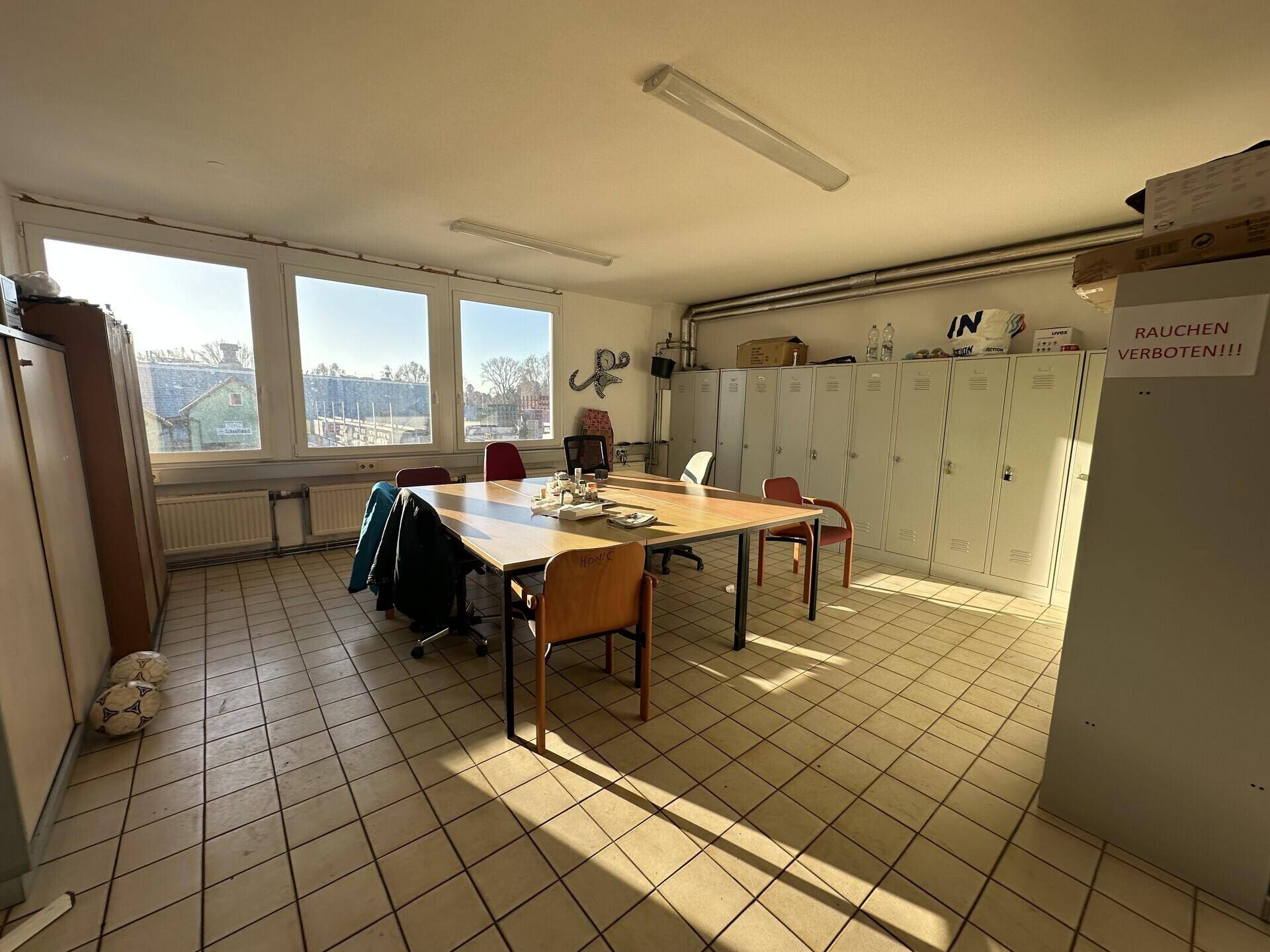 Pausenraum mit Tisch, Stühlen, Spinden und großen Fenstern, die viel Licht hereinlassen.