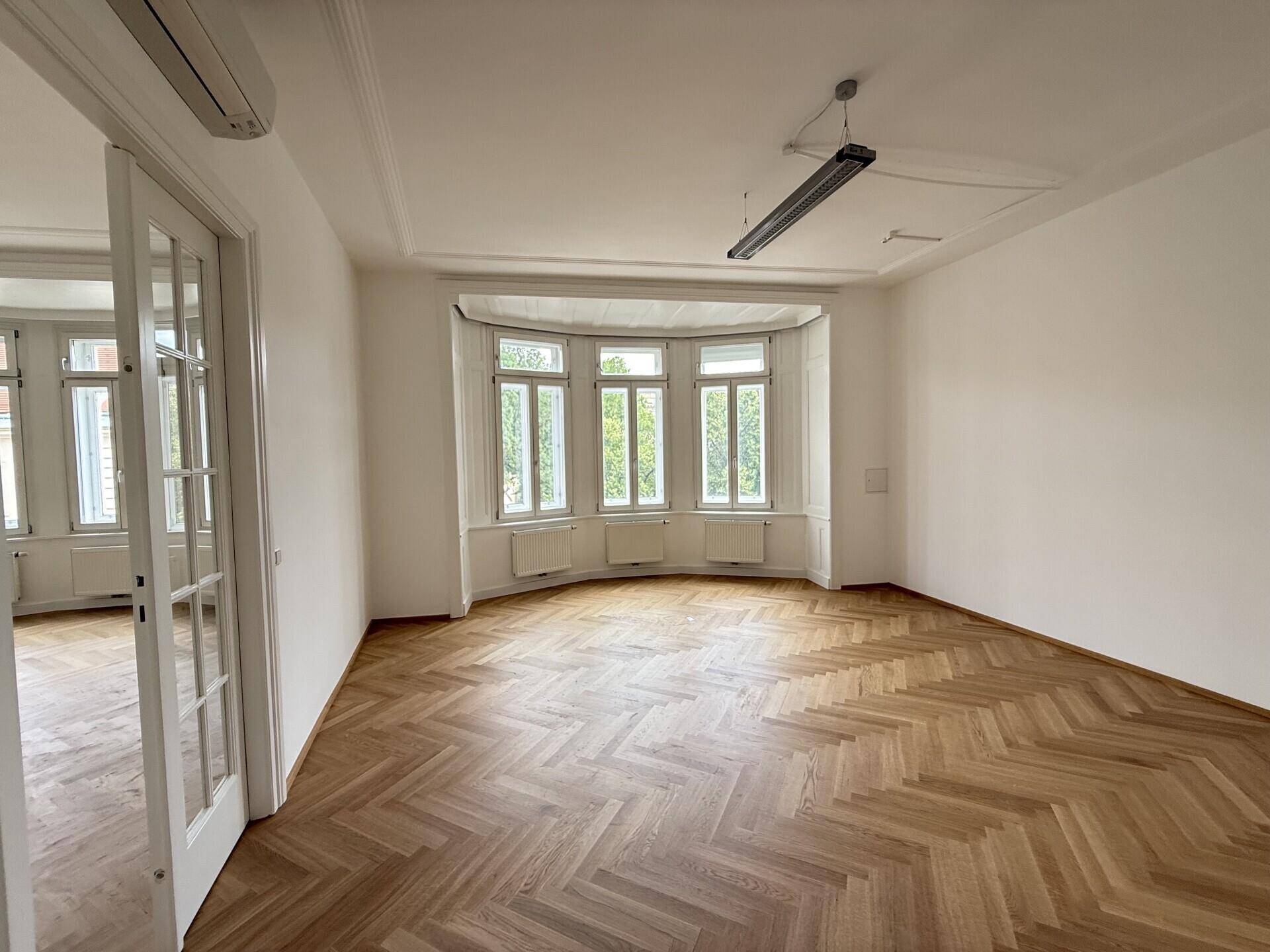 Geräumiges Büro mit Erkerfenstern, Fischgrätparkett und einer modernen Deckenleuchte, sehr hell.