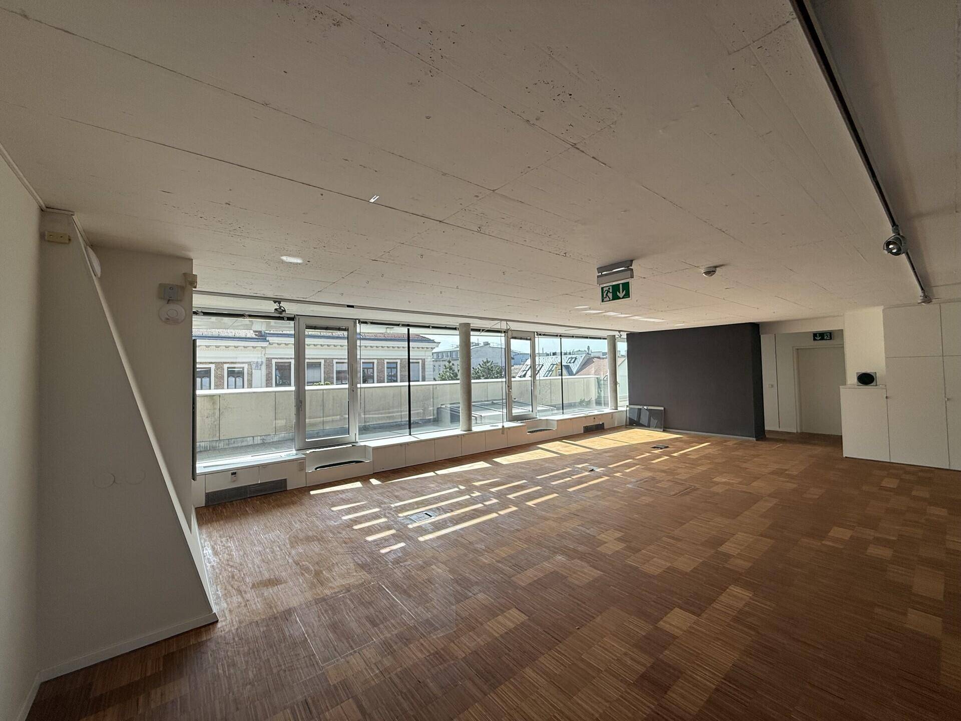 Großer, heller Büroraum mit Holzboden, Sichtbetondecke und einer Fensterfront mit Stadtblick.
