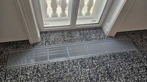 Detailaufnahme eines Heizgitters im Boden vor einem Fenster mit klassischem Balustraden-Geländer.