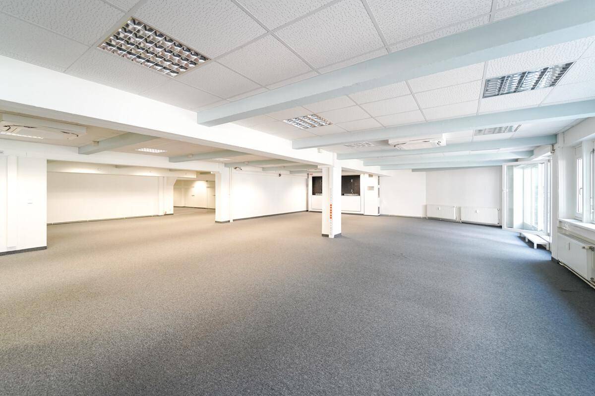 Weitläufiger Bürobereich mit grauem Teppichboden, Säulen und sichtbaren Deckenbalken, gut beleuchtet.