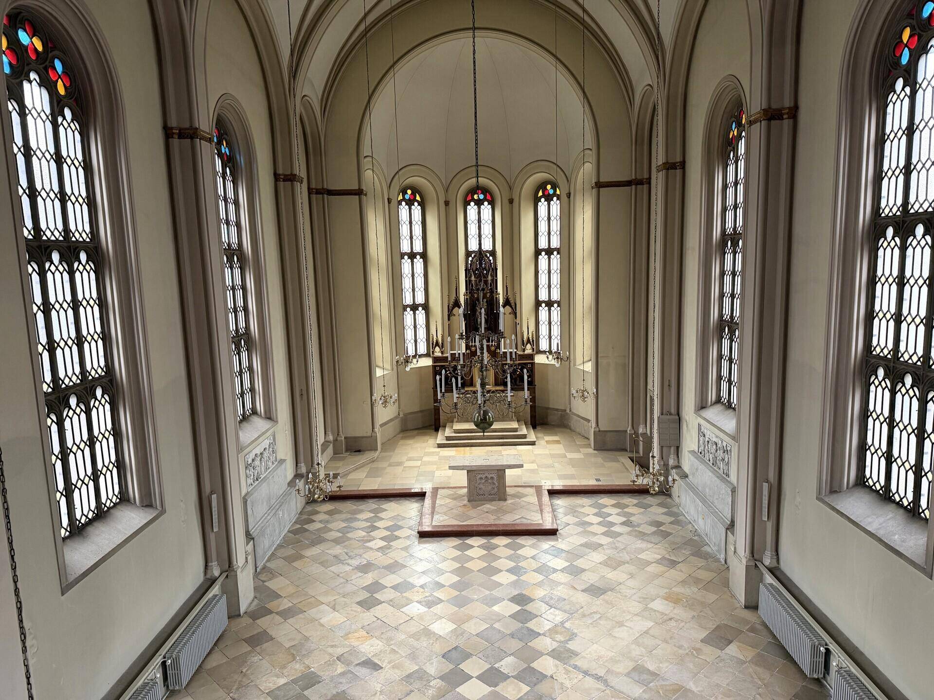 Blick von der Empore auf den Hauptsaal mit hohen Buntglasfenstern, Altarbereich und gemustertem Fliesenboden.