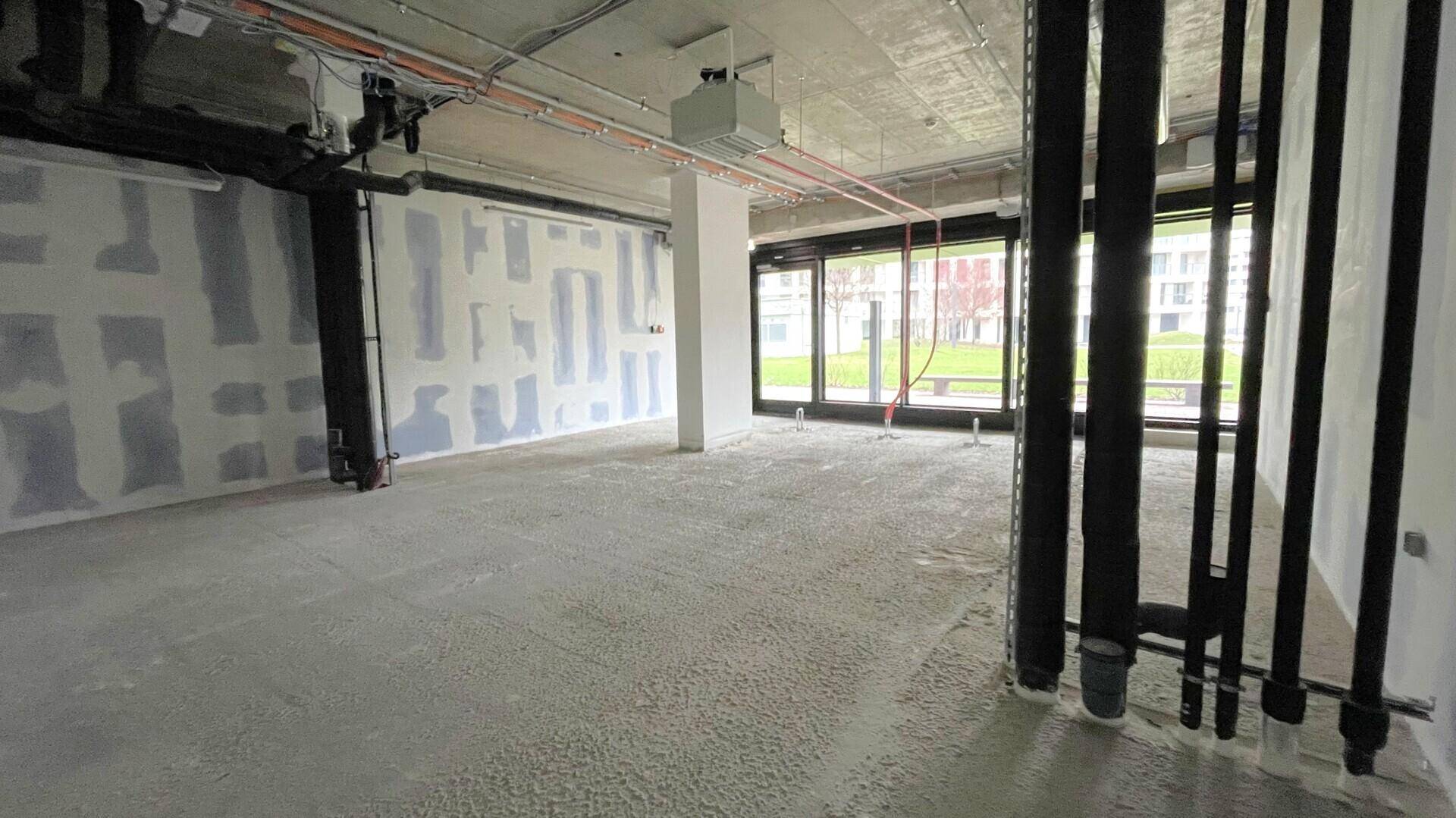 Geräumiger Gewerberaum im Rohbau mit sichtbaren Installationen, Betonboden und großen Fenstern, die den Außenbereich zeigen.