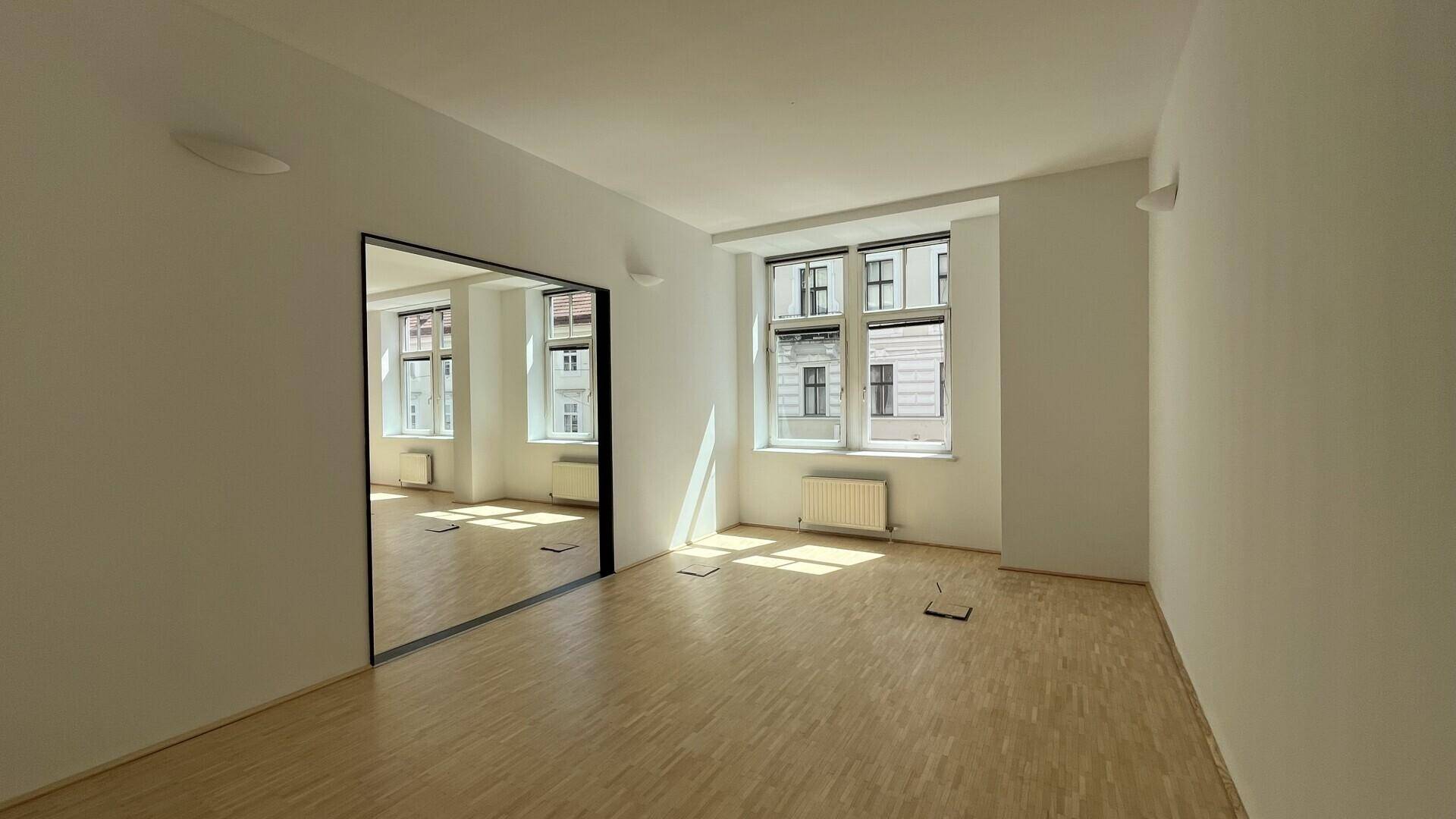 Leerer Büroraum mit hellem Holzfußboden, großen Fenstern und einem Wandspiegel, der den Raum erweitert.