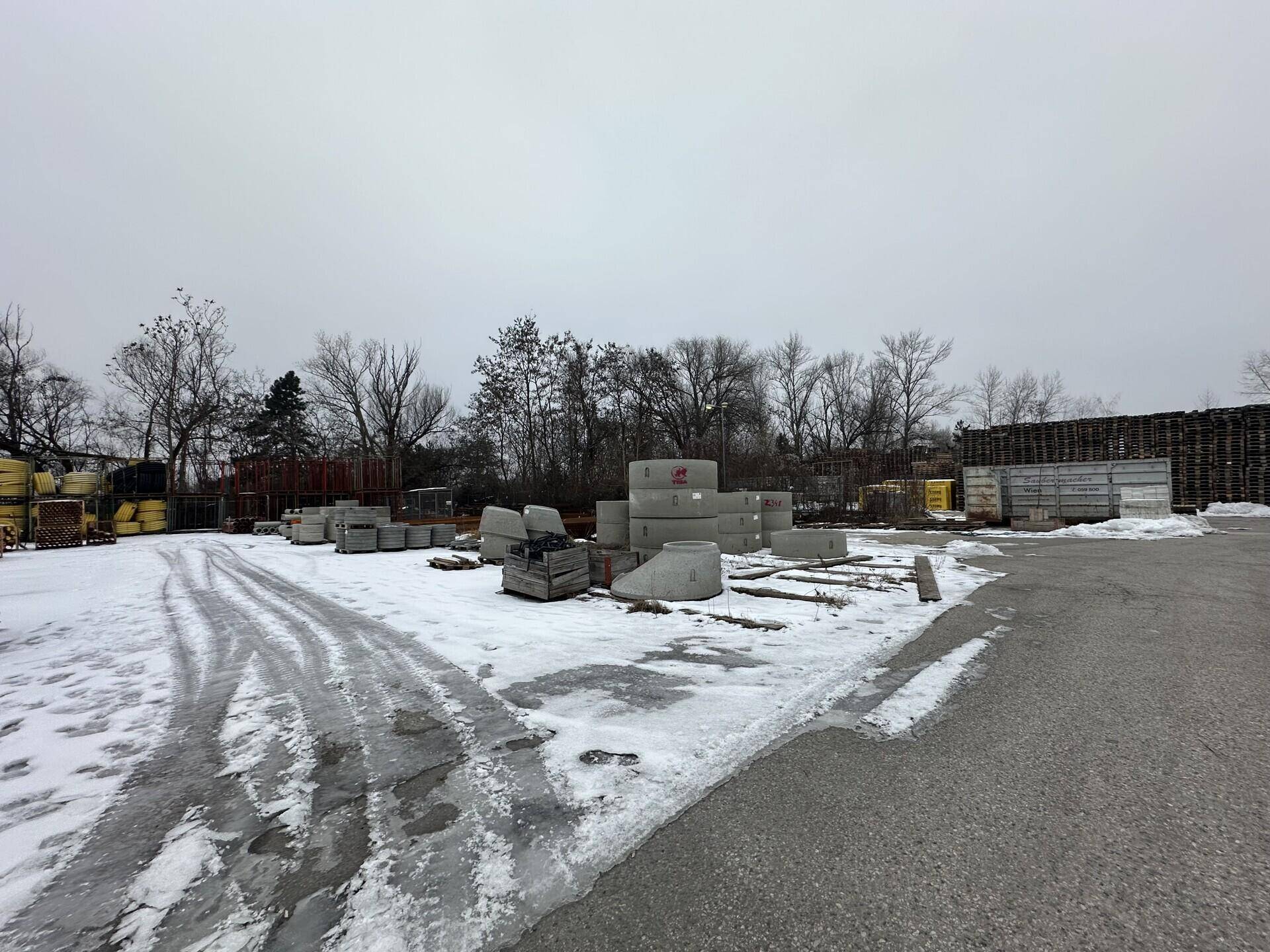 Außenlagerplatz mit verschiedenen Betonfertigteilen und Baumaterialien, bedeckt mit Schnee.
