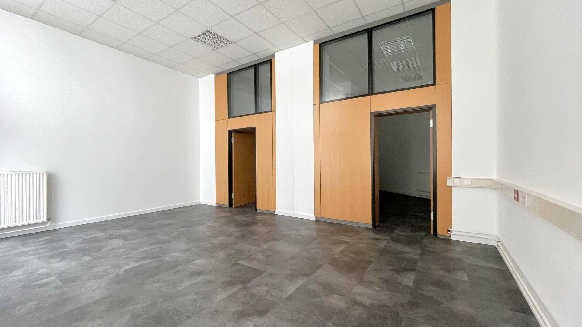 Leerer Büroraum mit dunklem Boden, weißen Wänden und hohen Fenstern über den Holztüren und Trennwänden.