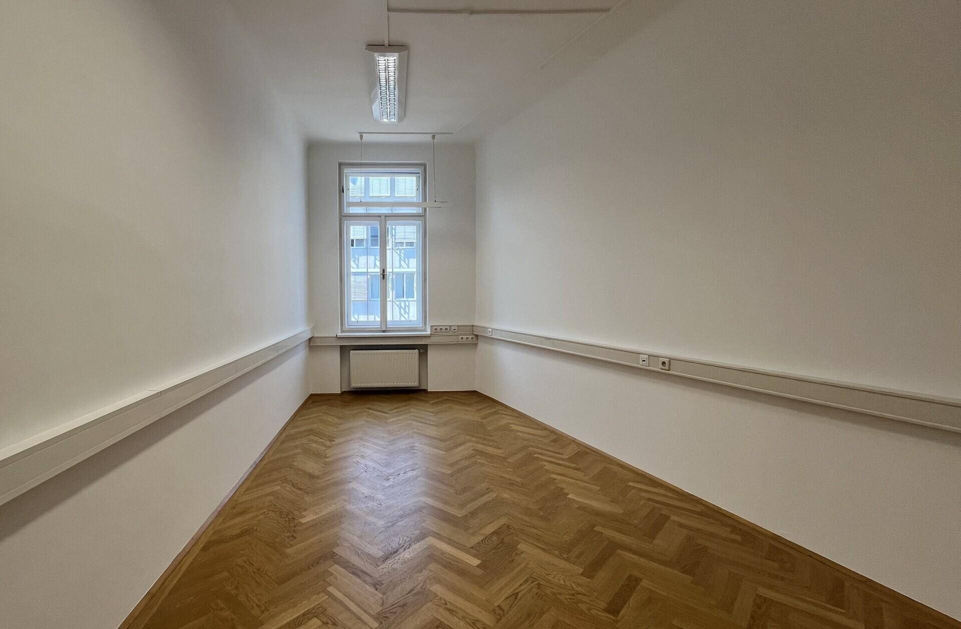 Schmaler Büroraum mit Fischgrätparkett, einem Fenster und Wandsteckdosenleisten, funktional gestaltet.
