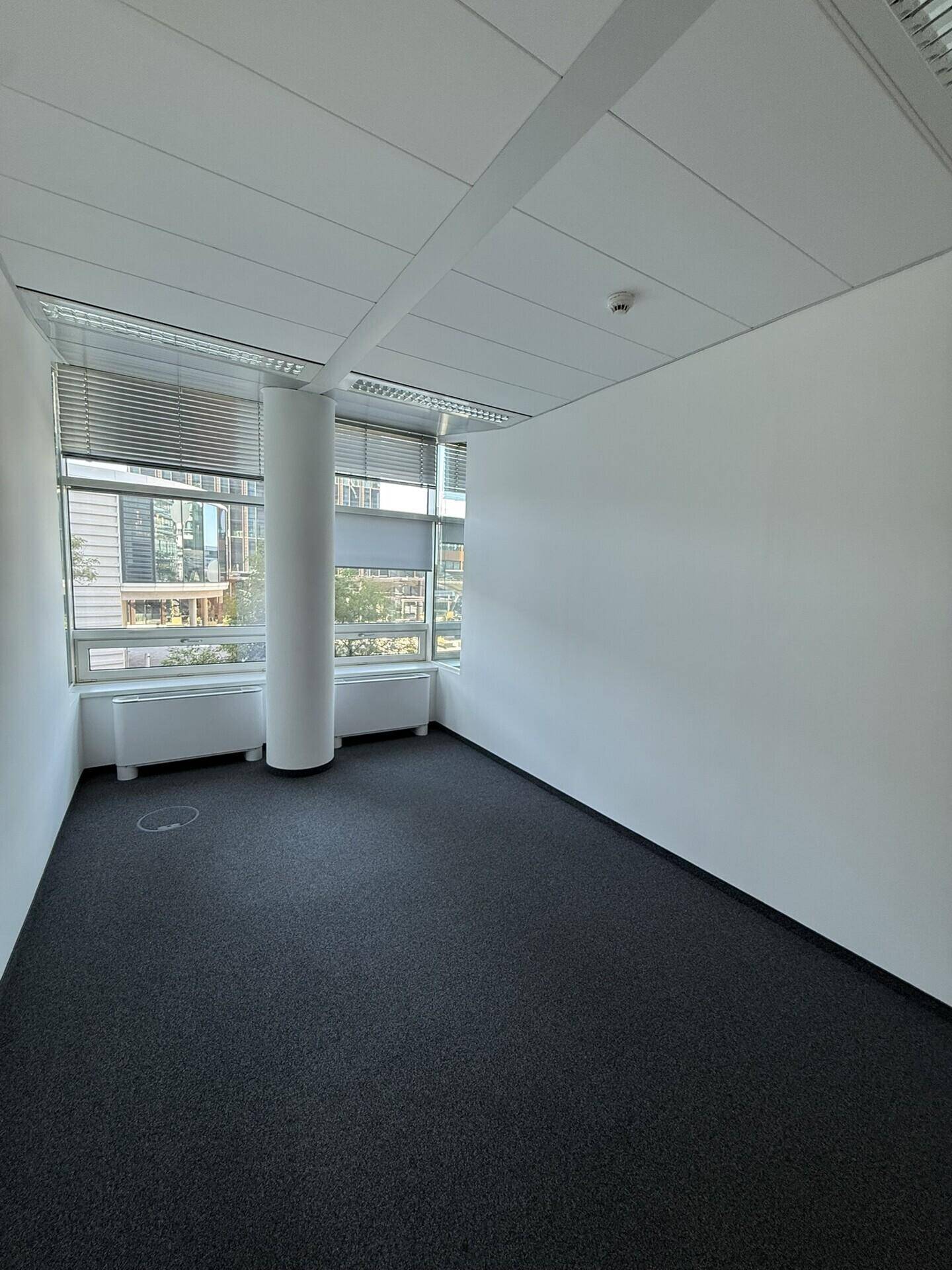 Leeres Büro mit Teppichboden, einer Säule und großen Fenstern, die den Raum mit natürlichem Licht füllen.