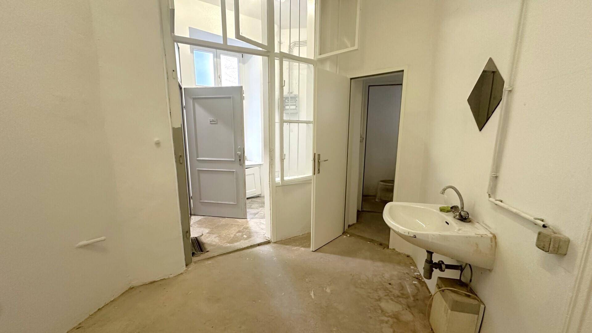 Ein einfacher Raum mit einem Waschbecken, einer Tür zu einem weiteren Bereich und einem Fenster mit Gitter.