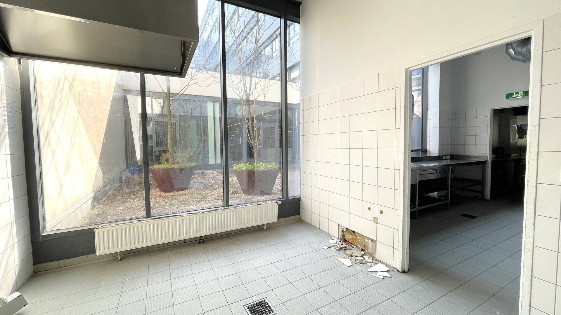 Gewerbeküche mit gefliesten Wänden, Blick auf den Innenhof durch große Fenster und Durchgang zu einem weiteren Raum.