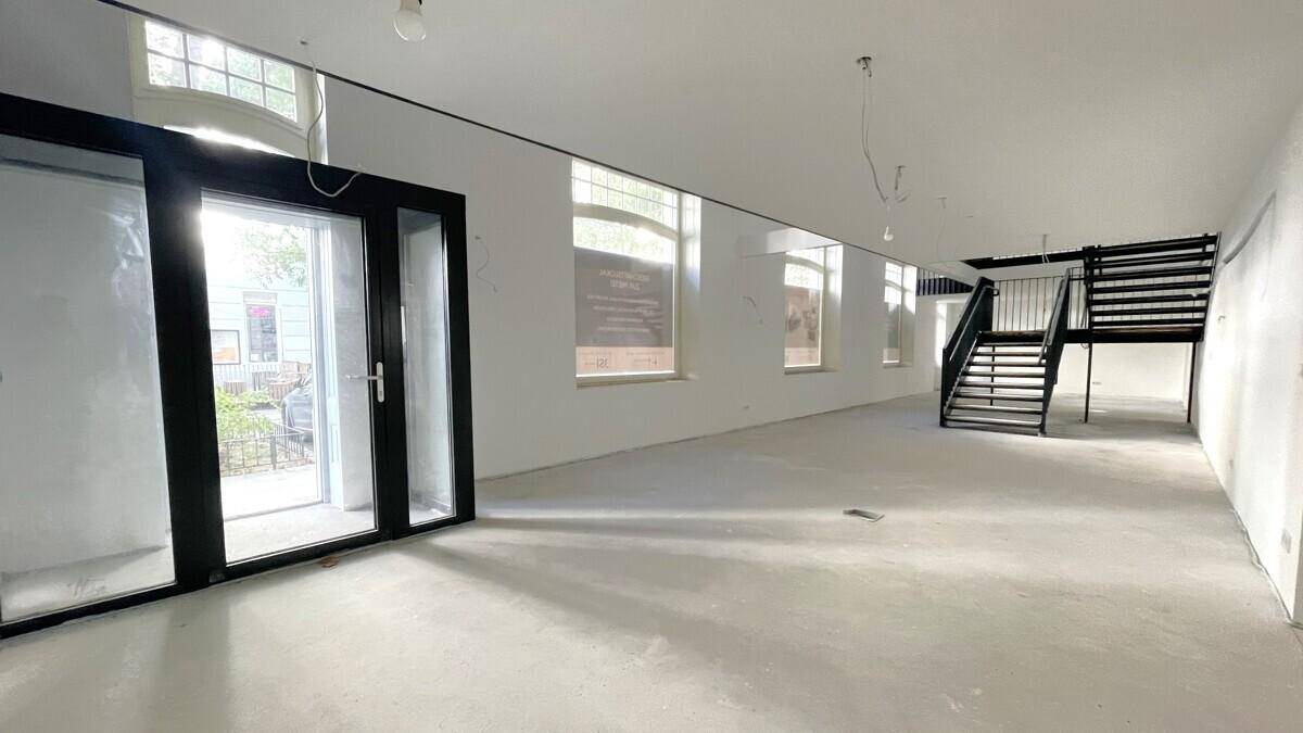 Großer, leerer Raum mit hohen Decken, großen Fenstern und einer schwarzen Treppe im Hintergrund.