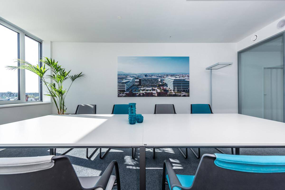 Heller Konferenzraum mit großem weißen Tisch, modernen Stühlen und Stadtansicht durch die Fenster.