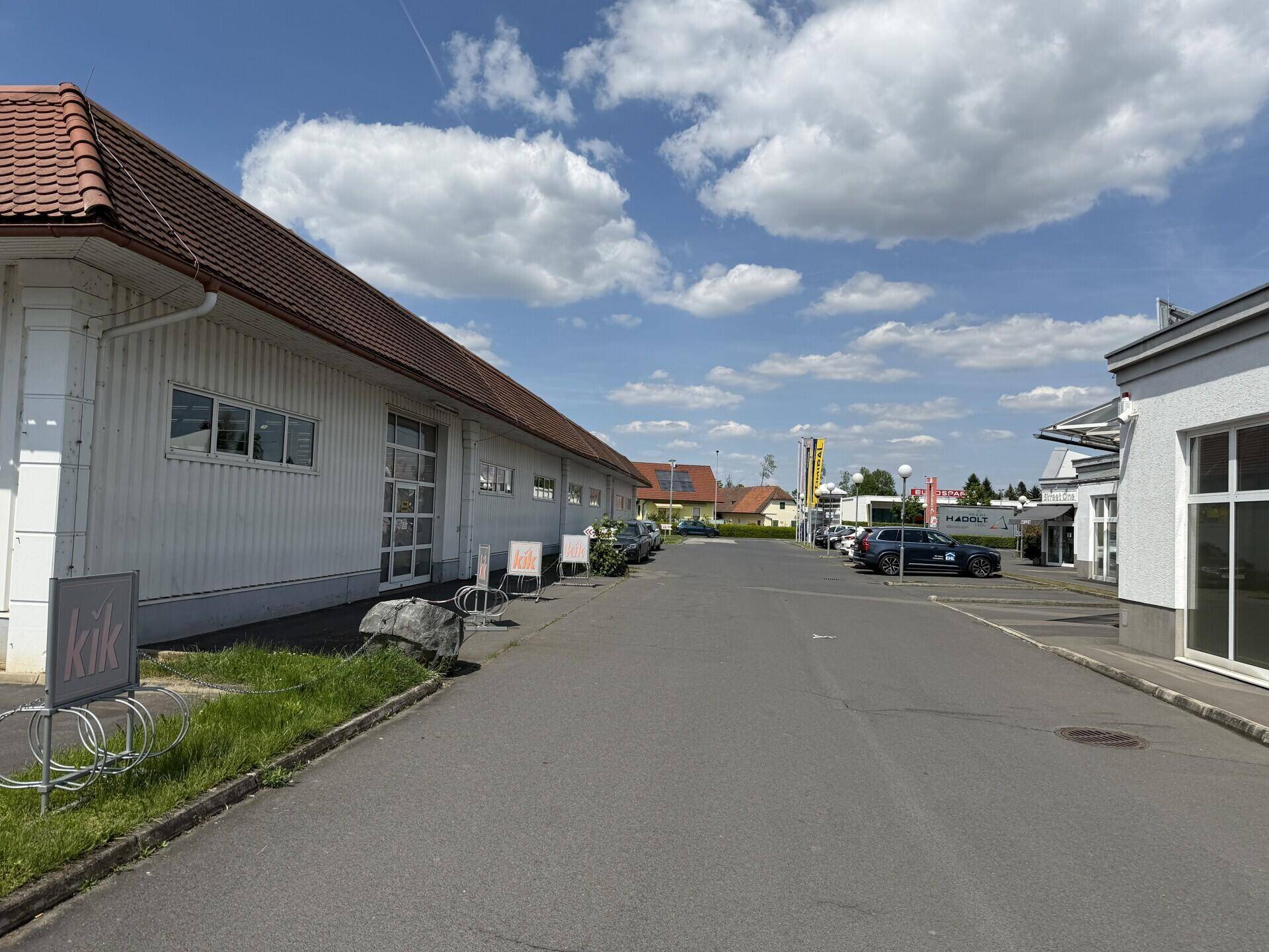 Zufahrtsweg zwischen Geschäftsgebäuden mit Parkplätzen und einem blauen Himmel mit weißen Wolken.