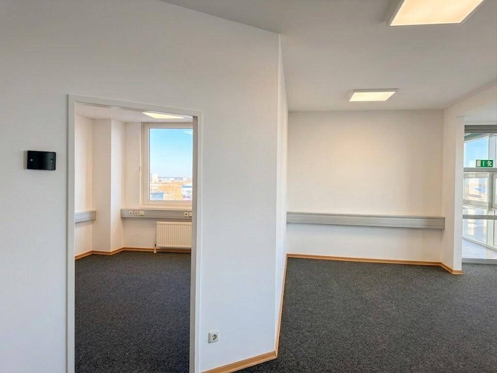Moderner Büroraum mit Teppichboden und weiß gestrichenen Wänden, der zu einem weiteren Zimmer führt.