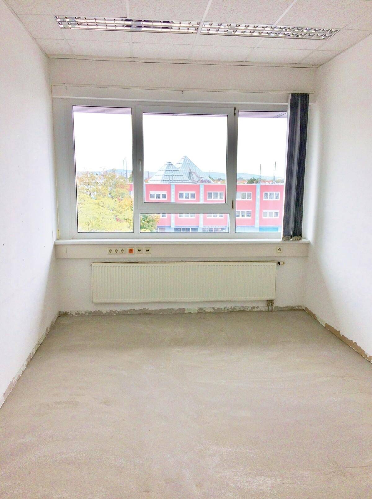 Kleinerer Büroraum mit Estrichboden, weißen Wänden und einem großen Fenster mit Blick auf andere Gebäude.