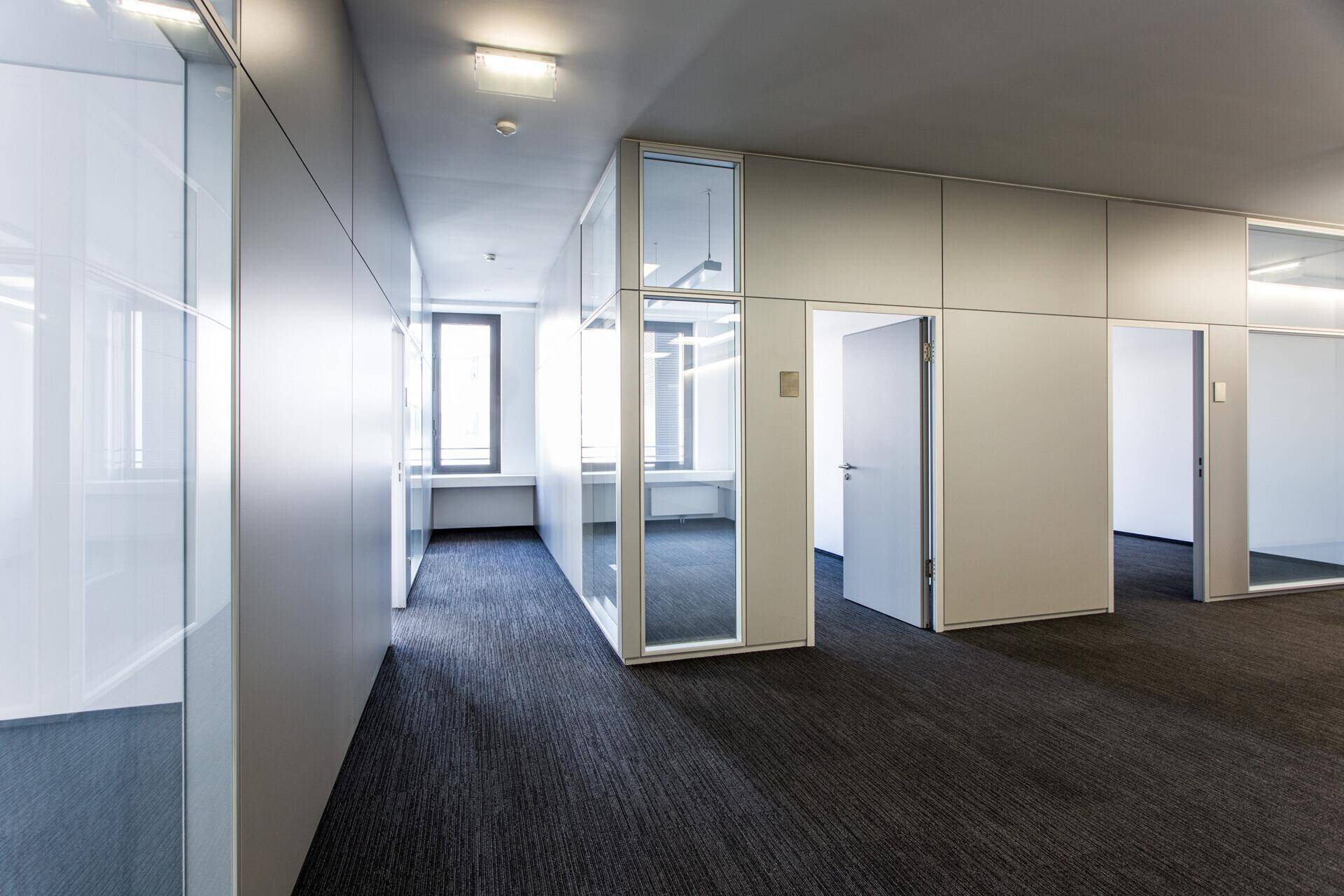 Moderner Bürogang mit Teppichboden und Glaswänden, der zu einzelnen Büros führt.