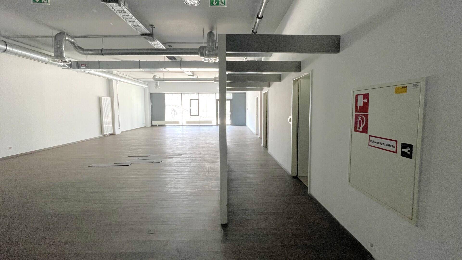 Langer, leerer Gewerberaum mit sichtbaren Deckeninstallationen und einem Durchgang zu weiteren Räumen.