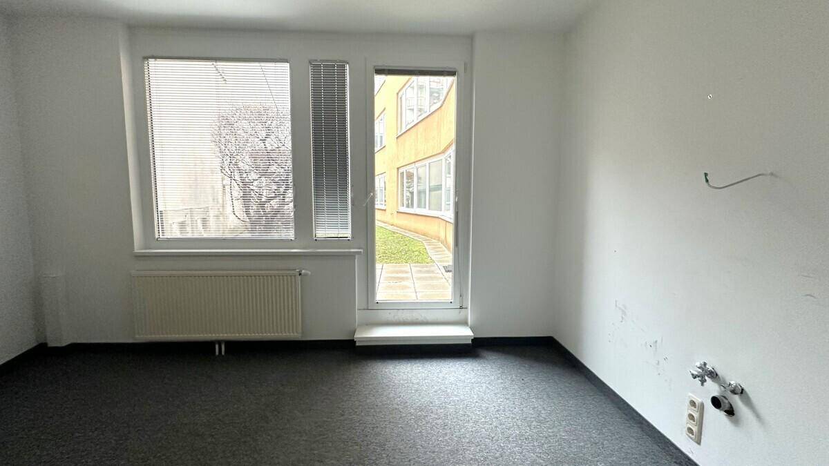 Leerer Büroraum mit Teppichboden und einer Glastür, die direkten Zugang zu einem Außenbereich ermöglicht.
