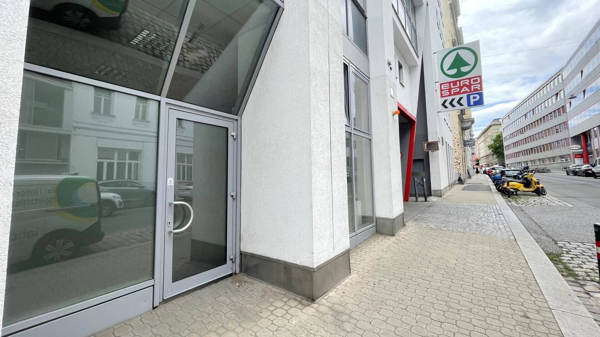 Eingangsbereich eines modernen Gebäudes mit Glasfassade und einem Supermarkt-Schild in der Nähe auf der Straße.