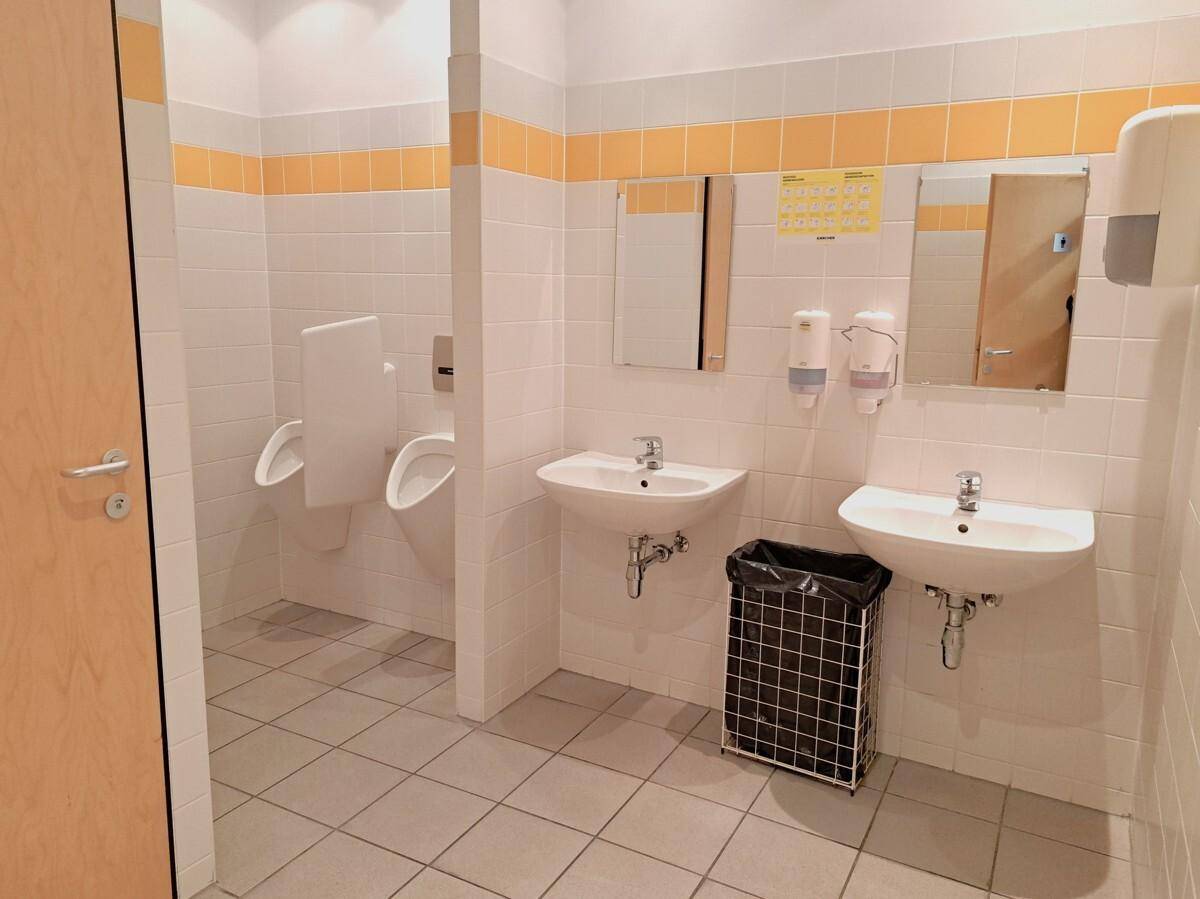 Sauberer öffentlicher Waschraum mit mehreren Urinalen, Waschbecken und weißen Fliesen an den Wänden.