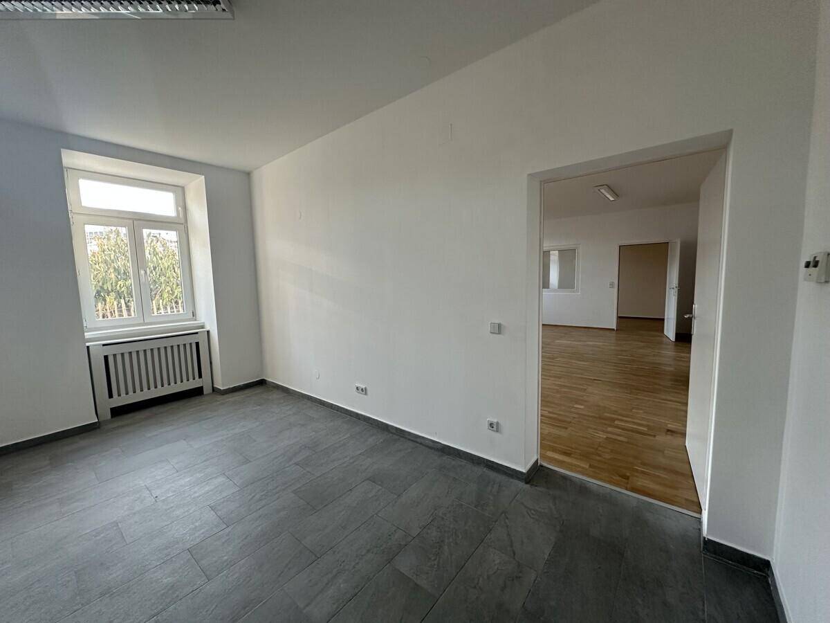 Heller Büroraum mit grauen Fliesen und einem Durchgang zu einem weiteren Raum mit Holzboden.