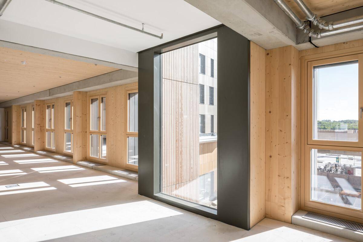 Langer, heller Flur mit Holzwänden, großen Fenstern und Blick auf die umliegenden Gebäude und den Himmel.