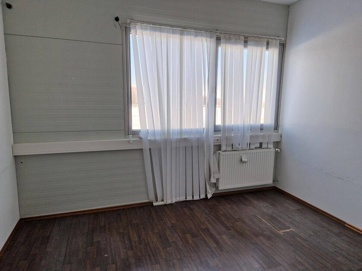 Leerer Raum mit dunklem Laminatboden, einer Wand mit großem Fenster, vertikalen Jalousien und Heizkörper.