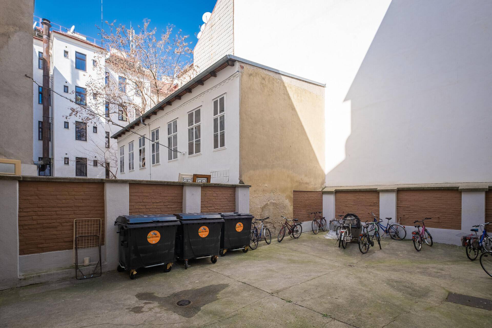 Hinterhofbereich mit Mülltonnen und vielen Fahrrädern, umgeben von Gebäudewänden und einem weiteren Gebäude.