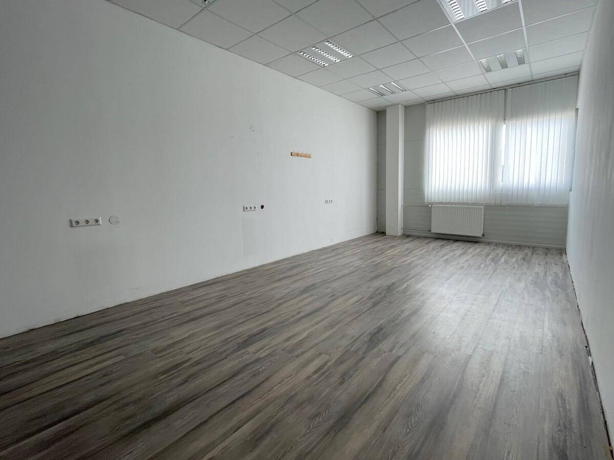 Leerer Büroraum mit Laminatboden, weißen Wänden und einem Fenster mit Jalousien.