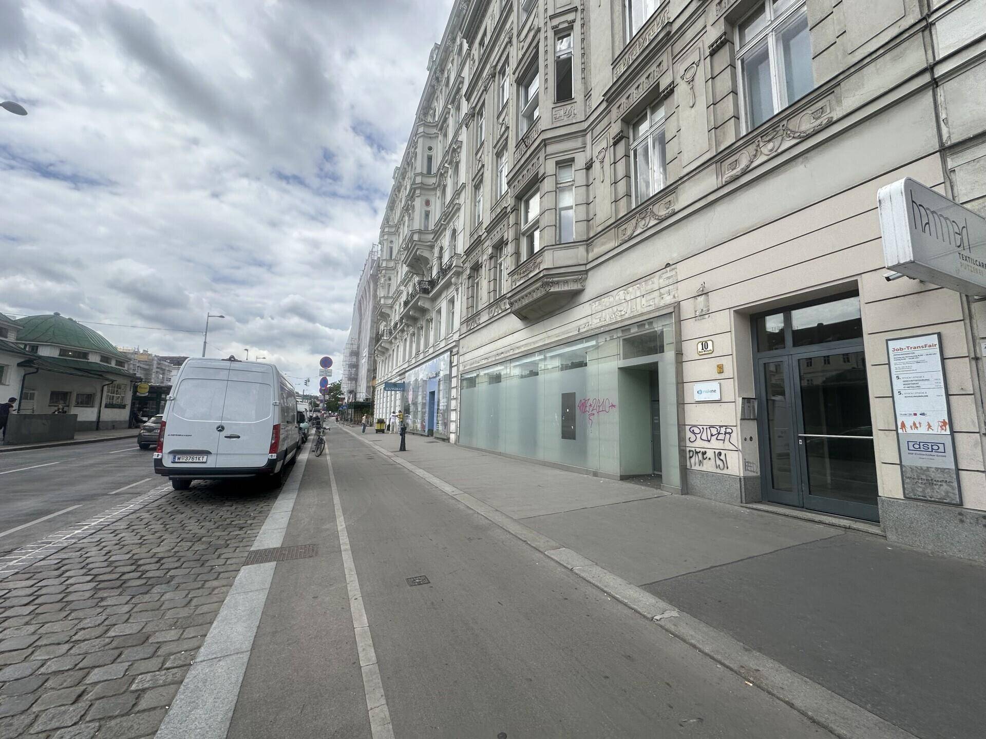 Blick entlang der Straße auf die historische Gebäudefassade mit Geschäften im Erdgeschoss und Passanten.