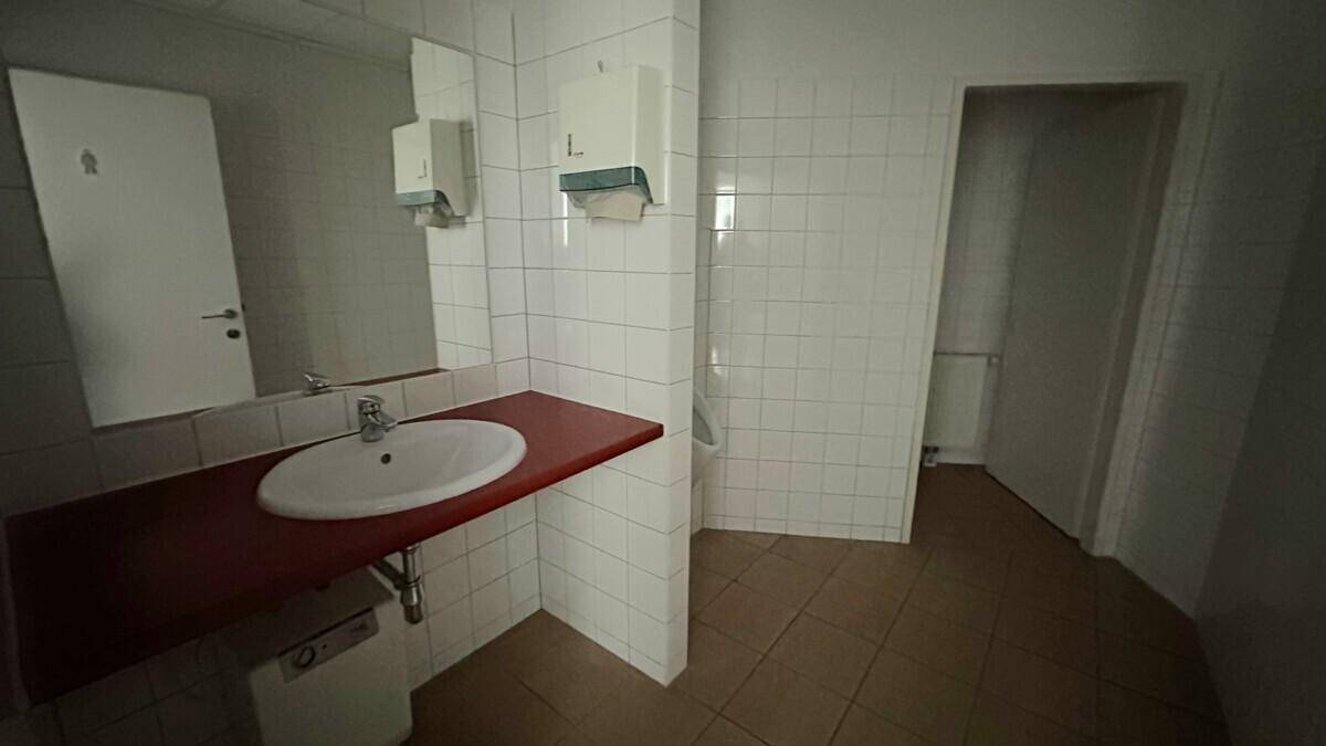Einfacher Toilettenraum mit weißen Fliesen, Waschbecken und roter Ablagefläche, gut beleuchtet.