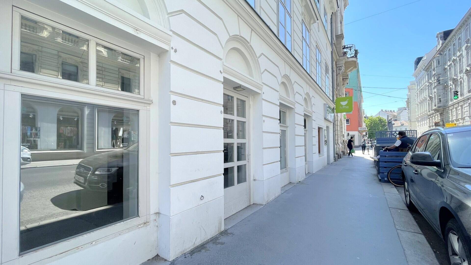 Seitenansicht eines klassischen Gebäudes an einer Straße mit großen Fenstern und einem breiten Bürgersteig.