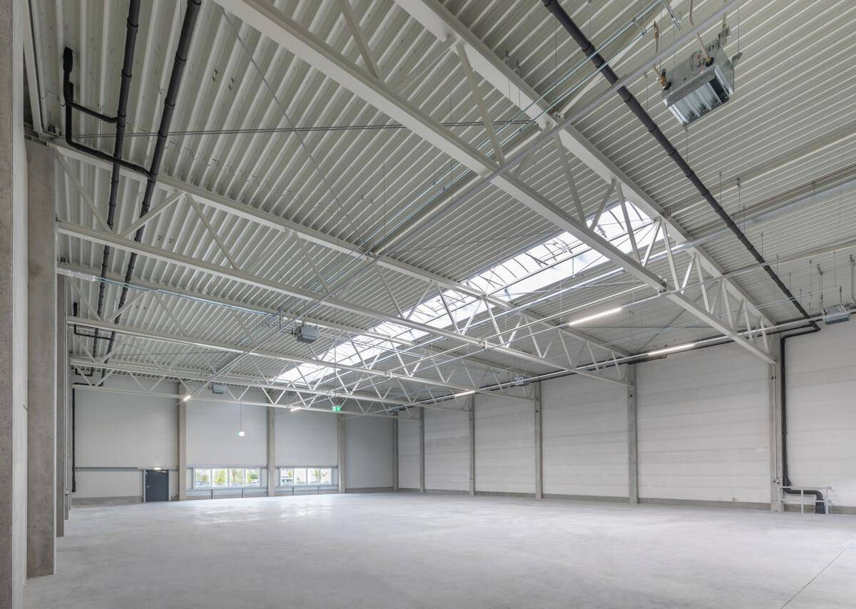 Geräumige, leere Lagerhalle mit hoher Decke, sichtbaren Stahlträgern und Oberlichtern für Tageslicht.